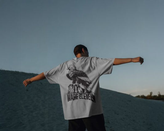 Person wearing a white t-shirt with a graphic design and text, standing on a sand dune with arms outstretched.